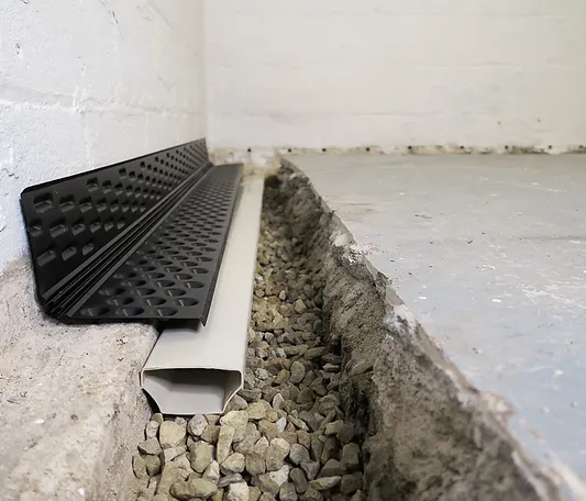 Close-up of an interior basement waterproofing drain installed along the wall-floor joint, with a dimpled wall flange, drainage channel, and gravel bed.